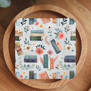 May include: Square coaster with a pale blue background featuring a pattern of books and flowers in shades of pink, orange, and green. The books are in various sizes and colours.