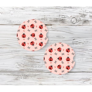 May include: Two round car coasters with a pink background. Each coaster features a repeating pattern of red ladybugs and black bows. The coasters are designed to fit in a car's cup holder and protect the interior from spills.