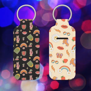 Op de afbeelding: Twee sleutelhangers met metalen ringen. Eén is zwart met een patroon van vredestekens, regenbogen, lippen en kersen. De andere is beige met een patroon van rolschaatsen, regenbogen en bliksemschichten. Beide hebben een retro, groovy esthetiek.