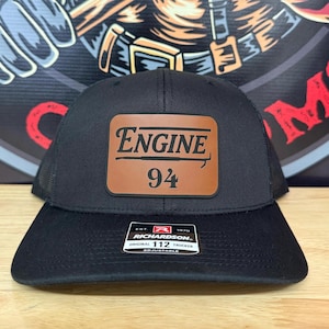May include: Black trucker hat with a brown leather patch. The patch reads "ENGINE" above the number "94". The hat has a mesh back and a curved peak. The Richardson brand label is visible.