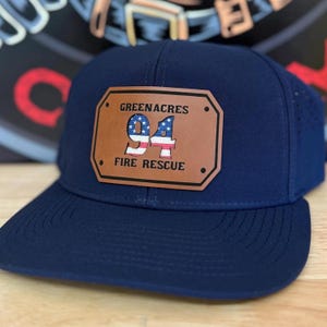 May include: A navy blue baseball cap with a brown leather patch. The patch features the text "GREENACRES 94 FIRE RESCUE" and a graphic of the American flag. The cap has a curved brim and a structured crown.