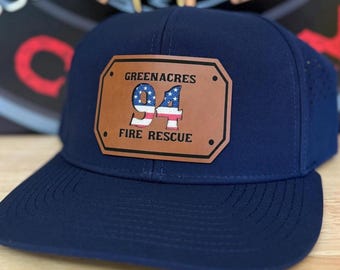 Könnte beinhalten: Marineblaue Baseballkappe mit einem braunen Lederaufnäher. Der Aufnäher zeigt den Text "GREENACRES 94 FIRE RESCUE" und eine Grafik der amerikanischen Flagge. Die Kappe hat einen gebogenen Schirm und eine strukturierte Krone.