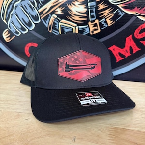 Peut inclure: Casquette de camionneur noire avec un motif rouge et blanc d'une hache de pompier et du drapeau américain. La casquette a une visière incurvée et un dos en filet. La casquette est réglable et porte une étiquette Richardson 112.