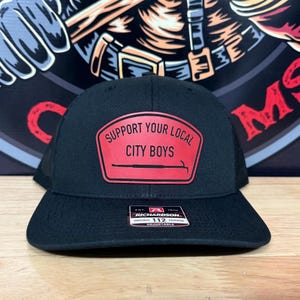 Op de afbeelding: Zwarte truckerpet met een rode patch met de tekst "SUPPORT YOUR LOCAL CITY BOYS". De pet heeft een gebogen klep en een verstelbare band. Het merk is Richardson 112.