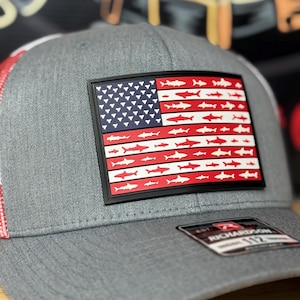Puede incluir: Gorra de béisbol gris con la parte trasera de malla roja y blanca. La parte delantera presenta un rectángulo negro con un diseño de la bandera estadounidense, pero con siluetas de tiburones en lugar de estrellas y rayas. La marca "RICHARDSON 112" es visible en una etiqueta.
