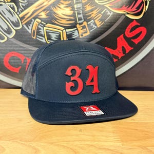 May include: A navy blue snapback hat with a mesh back and a flat bill. The front features the number 34 in raised red lettering. A rectangular sticker on the bill displays the Richardson logo with the text FLAT BILL SNAPBACK EST 1970.