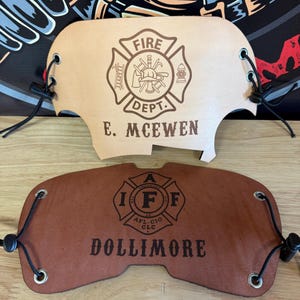 Puede incluir: Dos protectores de casco de bombero de cuero. El protector superior es de color canela con las palabras "FIRE DEPT." y "E. MCEWEN" debajo de un emblema de bombero. El protector inferior es marrón con las palabras "DOLLIMORE" y un emblema de bombero.
