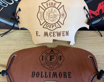May include: Two leather firefighter helmet shields. The top shield is tan with the words "FIRE DEPT." and "E. MCEWEN" below a fire department emblem. The bottom shield is brown with the words "DOLLIMORE" and a fire department emblem.