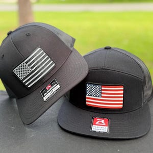 May include: Two black flat bill snapback hats with American flag patches. One patch features a black and white flag, the other a red, white, and blue flag. Both hats have a mesh back and a Richardson 112 tag.