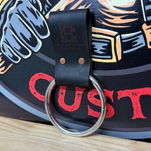 May include: A black leather belt loop strap with two copper rivets and a silver metal ring. The strap is embossed with the text LEATHERHEAD CUSTOMS and a logo. It is attached to a black surface featuring a graphic illustration with red text.