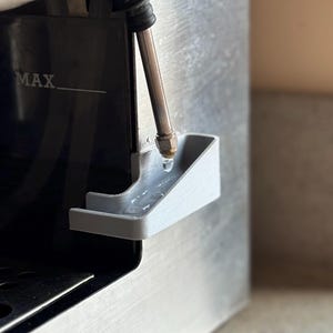 May include: A close-up of a silver coffee machine with a black water tank labelled "MAX". A silver metal tube with a black rubber hose is connected to a grey plastic drip tray.