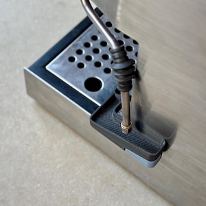 Puede incluir: Primer plano de un componente de máquina de espresso de acero inoxidable. La imagen muestra una varilla de vapor, una bandeja de goteo con agujeros circulares y un pequeño componente rectangular negro con un borde gris.