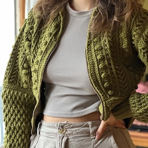 Op de afbeelding: Een groene gehaakte bomberjack met een ritssluiting. De jas heeft een textuur met reliëfknopjes.