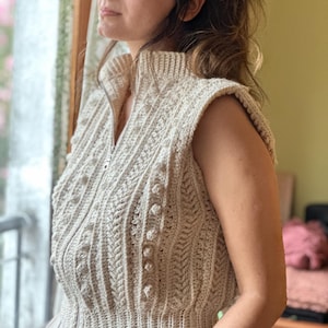 May include: A cream-colored crocheted vest with a zipper closure and a cable knit pattern. The vest has a high neckline and is sleeveless.
