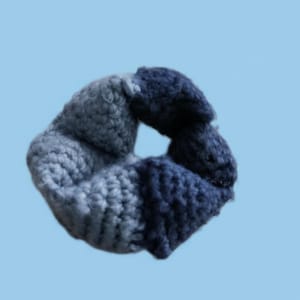 Könnte beinhalten: Ein handgefertigtes, gehäkeltes Haargummi in Blau- und Grautönen. Das Haargummi ist aus einem dicken, strukturierten Garn gefertigt und soll das Haar in einem Pferdeschwanz oder Dutt halten. Der Hintergrund ist ein helles Blau.