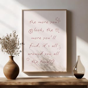 May include: A framed print with the handwritten text "the more you look, the more you'll find. it's all around you all the time" in a soft red hue. The frame is a light wood color. A brown vase with dried flowers and a decorative glass bottle are on a wooden surface.