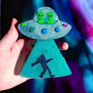 Puede incluir: Un jabón triangular azul verdoso con una silueta negra de un extraterrestre caminando. La parte superior del jabón es un OVNI transparente y esmerilado con dos extraterrestres verdes en su interior.