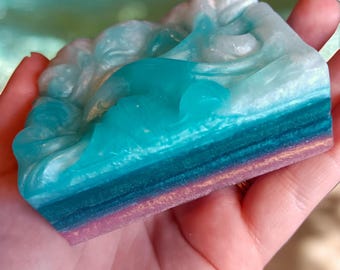 May include: A handmade soap bar with a blue and white swirl design. The soap has a layered look with a pink, green, and gold shimmer.