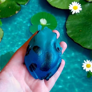 May include: A blue frog figurine with black spots is held in a hand. The frog is set against a backdrop of water, lily pads, and daisies. The figurine has a glossy finish and is a vibrant shade of blue.