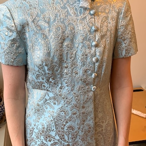 May include: A light blue dress with a silver and gold floral brocade pattern. The dress features short sleeves, a high collar, and a row of matching buttons down the front. A small bow detail is at the neckline.