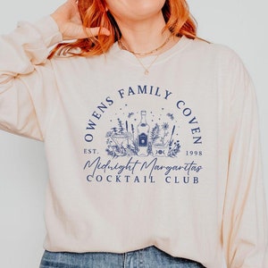 Könnte beinhalten: Ein cremefarbenes Langarmshirt mit einem blauen Grafikdesign, auf dem "Owens Family Coven Est. 1998 Midnight Margaritas Cocktail Club" steht.