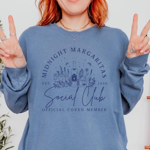 Könnte beinhalten: Ein blaues Langarmshirt mit einem Motiv einer Tequilaflasche, einem Margarita-Glas und floralen Elementen. Der Text "Midnight Margaritas Social Club Est. 1998 Official Coven Member" ist auf dem Shirt aufgedruckt.