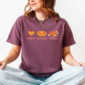 May include: A maroon t-shirt featuring a graphic with a heart, pumpkin pie, and turkey. The text "FAMILY DESSERT TURKEY" is printed below the images. The shirt is made of a soft material.