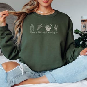 Puede incluir: Sudadera verde oscuro con el texto "there's a little witch in all of us" e ilustraciones botánicas. El diseño incluye un salero, ramitas de lavanda, un mortero y una maja, y otras hierbas. Un estilo casual y cómodo.