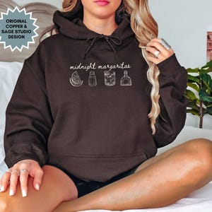 Könnte beinhalten: Ein dunkelbrauner Kapuzenpullover mit Kordelzug und einer weißen Grafik mit der Aufschrift "midnight margaritas" mit Illustrationen einer Limette, einer Flasche, einem Glas und einer Tequila-Flasche. Der Hoodie hat ein Etikett mit der Aufschrift "Original Copper & Sage Studio Design".