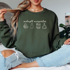Könnte beinhalten: Ein olivgrünes Sweatshirt mit dem weißen Schriftzug "midnight margaritas". Darunter befinden sich weiße Strichzeichnungen einer Limettenscheibe, eines Salzstreuers, eines Cocktails und einer Flasche. Das Sweatshirt hat einen Rundhalsausschnitt und lange Ärmel.
