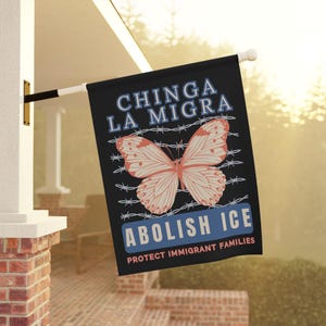May include: A black rectangular flag featuring a central peach-colored butterfly graphic overlaid on white barbed wire. The text reads CHINGA LA MIGRA in blue, ABOLISH ICE in white on a blue background, and PROTECT IMMIGRANT FAMILIES in peach text.