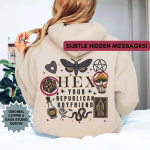 May include: Beige hoodie with a graphic design on the back. The design includes a moth, pentagram, mushrooms, and the text "HEX YOUR REPUBLICAN BOYFRIEND." The hoodie also features the text "SUBTLE HIDDEN MESSAGES!" and "ORIGINAL COPPER & SAGE STUDIO DESIGN."