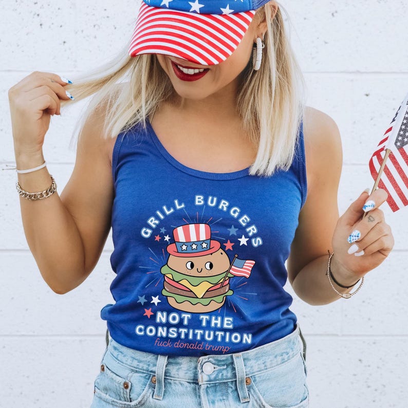 May include: A blue vest top featuring a cartoon burger wearing a patriotic hat and holding a small American flag. The text "GRILL BURGERS" and "NOT THE CONSTITUTION" are above and below the burger, with "fuck donald trump" below.