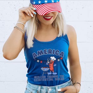 May include: A blue tank top with the word "AMERICA" in white arched text. Below is a cartoon hot dog grilling on a barbecue, with the text "GRILLING THROUGH THE EXISTENTIAL DREAD". The person is wearing a red, white, and blue baseball cap.