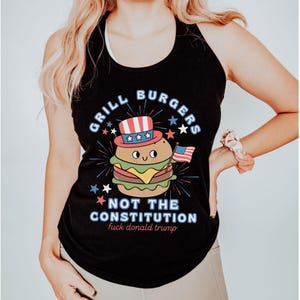 May include: Black tank top featuring a burger character in a patriotic hat and holding a small American flag. Text reads "Grill Burgers Not The Constitution" with additional text below.