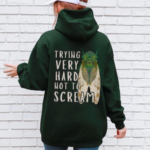 Könnte beinhalten: Dunkelgrüner Kapuzenpullover mit dem weißen Text "TRYING VERY HARD NOT TO SCREAM". Auf der Rückseite ist eine detaillierte Illustration einer Zikade mit grünen und beigen Flügeln aufgedruckt. Der Hoodie hat eine Kapuze und lange Ärmel.