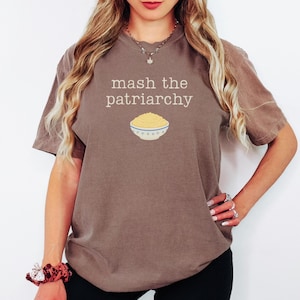 May include: A brown t-shirt featuring the phrase "mash the patriarchy" and an image of mashed potatoes in a bowl. The shirt has short sleeves and a solid color.