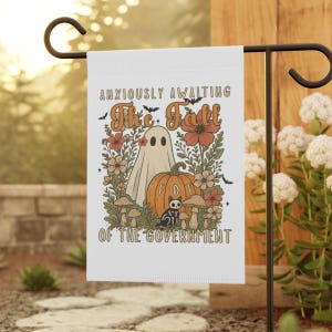 Op de afbeelding: Witte tuinvaandel met een grillig ontwerp met een spook, pompoen, bloemen en paddenstoelen. De tekst luidt "Anxiously Awaiting The Fall Of The Government". De vlag wordt buiten op een zwarte metalen standaard getoond.