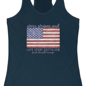 May include: Navy blue tank top with a racerback design. The front features a distressed American flag graphic with the text "stars stripes and" above and "LATE STAGE CAPITALISM fuck donald trump" below the flag.