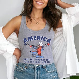 May include: A gray tank top with the word "AMERICA" in blue arched text. Below the text is a cartoon hot dog grilling on a barbecue. The text "GRILLING THROUGH THE EXISTENTIAL DREAD" is below the cartoon.