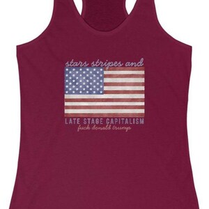 May include: Burgundy racerback tank top with a graphic of the American flag and the text "stars stripes and LATE STAGE CAPITALISM fuck donald trump" in a distressed font. The top has a scoop neck and a racerback design.