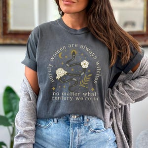 May include: A dark gray t-shirt with a graphic print of a snake with flowers and stars. The text on the shirt reads "Unruly women are always witches no matter what century we're in".