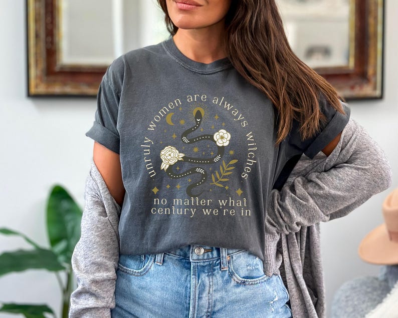 May include: A dark gray t-shirt with a graphic print of a snake with flowers and stars. The text on the shirt reads "Unruly women are always witches no matter what century we're in".