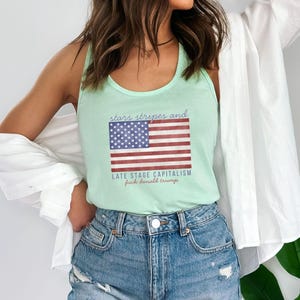 May include: A light green tank top with the American flag graphic and the text "stars stripes and late stage capitalism fuck donald trump". The tank top is paired with denim shorts.