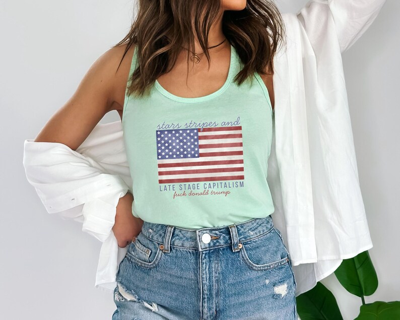 May include: A light green tank top with the American flag graphic and the text "stars stripes and late stage capitalism fuck donald trump". The tank top is paired with denim shorts.