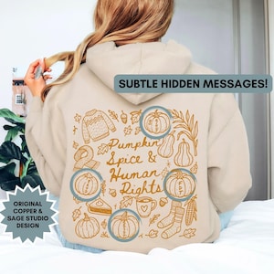 May include: Beige hoodie with a fall-themed graphic on the back. The design includes pumpkins, leaves, a sweater, and the text "Pumpkin Spice & Human Rights." The hoodie also has the text "Subtle Hidden Messages!" and "Original Copper & Sage Studio Design."