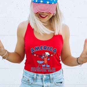 May include: Red tank top with the word "AMERICA" in blue and white lettering. The graphic features a cartoon hot dog grilling on a barbecue. The text "GRILLING THROUGH THE EXISTENTIAL DREAD" is below the graphic. The person is wearing a red, white, and blue baseball cap.