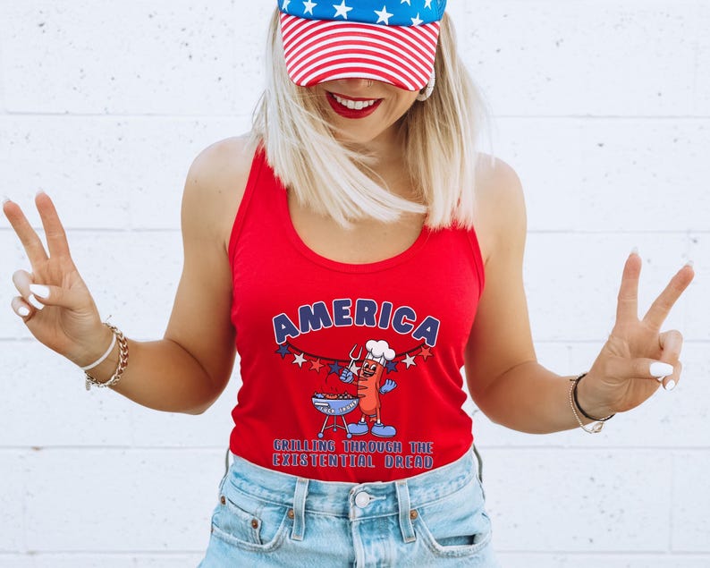 May include: Red tank top with the word "AMERICA" in blue and white lettering. The graphic features a cartoon hot dog grilling on a barbecue. The text "GRILLING THROUGH THE EXISTENTIAL DREAD" is below the graphic. The person is wearing a red, white, and blue baseball cap.