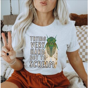 May include: Light gray t-shirt with the text "TRYING VERY HARD NOT TO SCREAM" and a green and tan cicada illustration. The shirt is a crew neck style.
