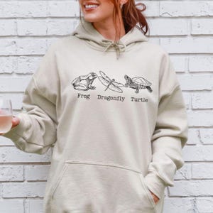 May include: A cream-colored hoodie with a frog, dragonfly, and turtle graphic. The text "Frog Dragonfly Turtle" is printed below the graphic.